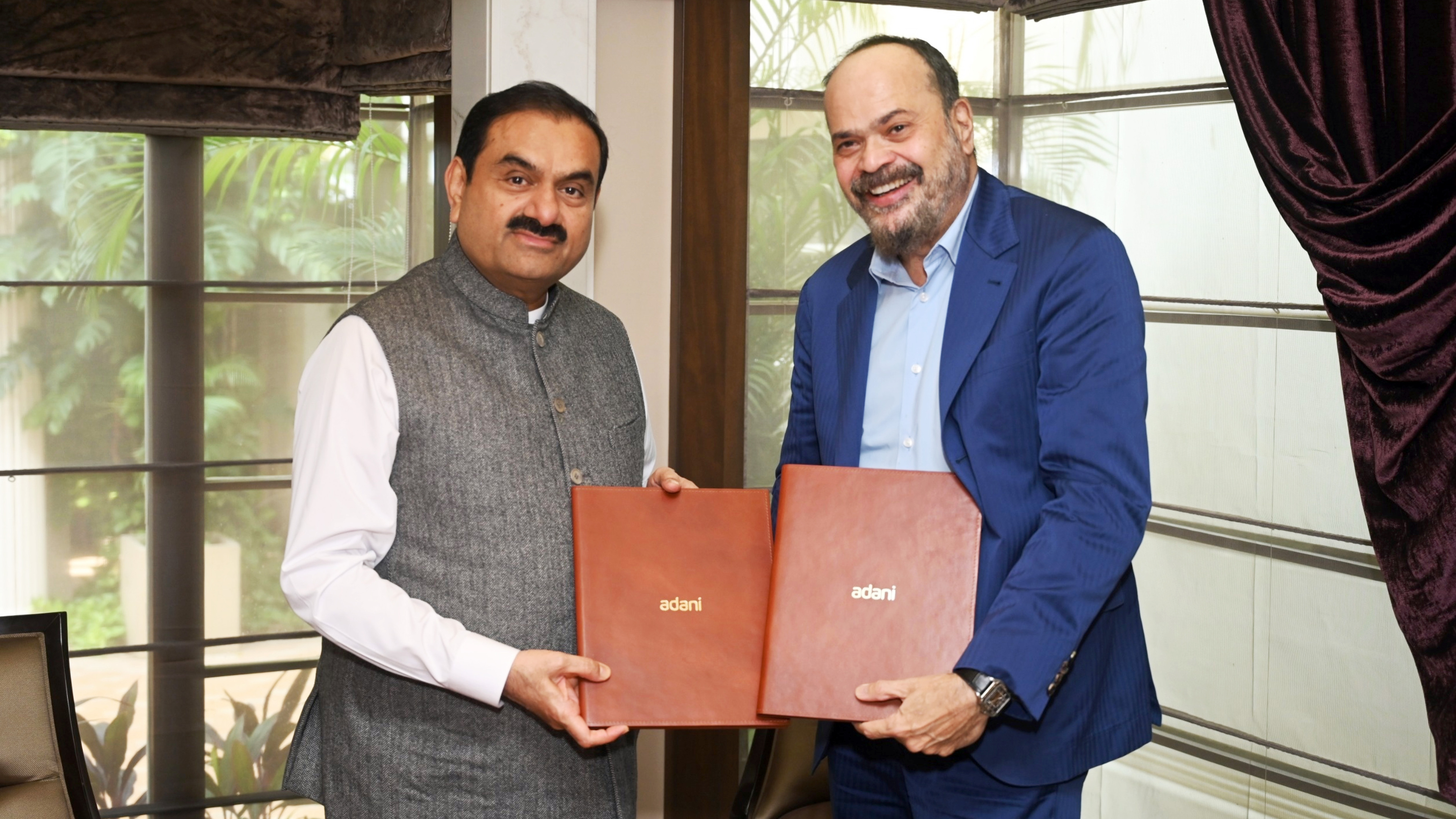 Two men, one in a vest and one in a suit, smile while holding "Adani" branded folders, signifying an agreement.