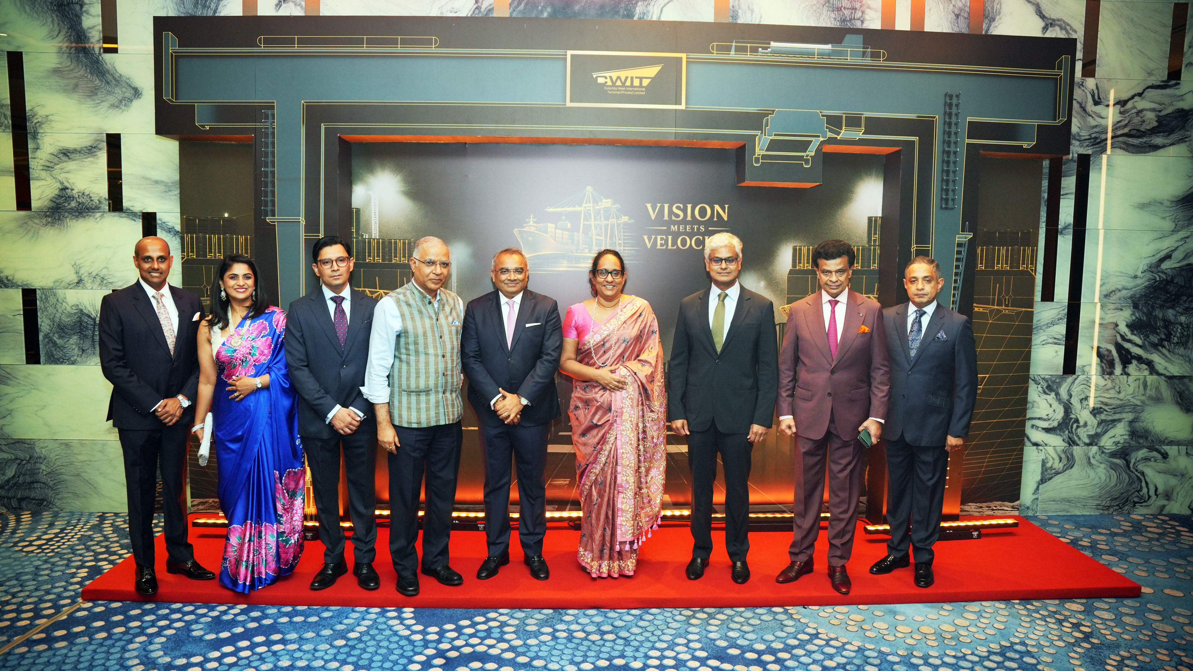 Nine people, including men in suits and women in saris, stand formally on a red carpet in front of a stage.