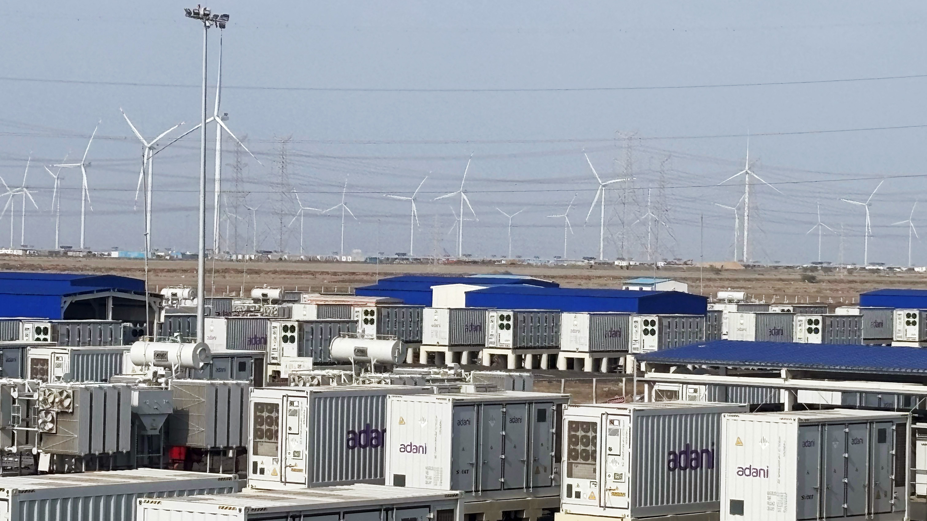Adani wind power and energy storage facility with numerous containers and wind turbines under a clear sky.