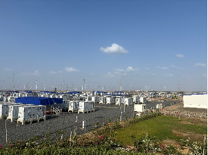 Rows of white container units at a large energy storage or data center facility, with wind turbines in the background.