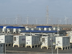 Numerous container-like structures in a field, with wind turbines and power lines on the horizon under a clear sky.