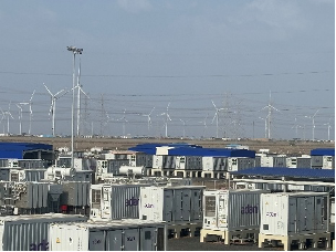 Energy storage containers in front of a wind farm under a clear sky.